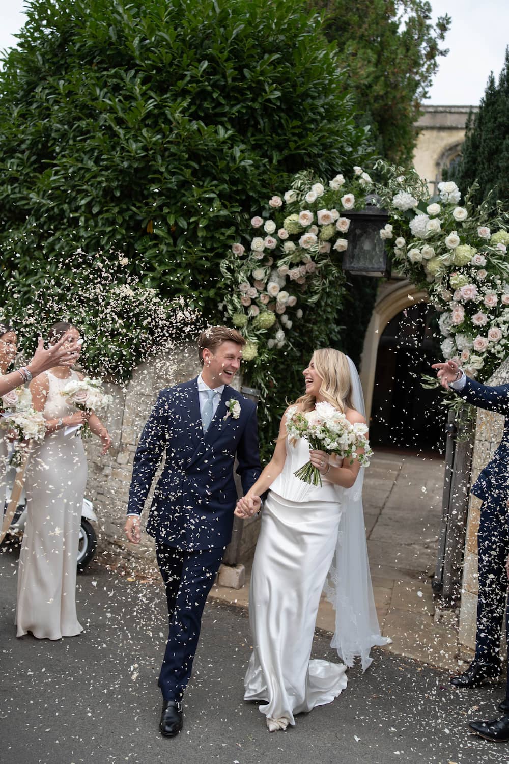 A bride in a white satin wedding gown and a groom in a navy suit walk hand-in-hand under a floral arch, with guests throwing flower petals. The bride holds a bouquet of white and green flowers, unedited.