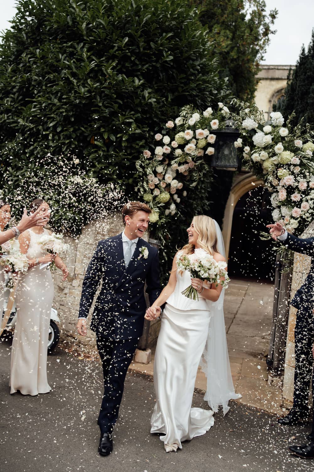 A bride in a white satin wedding gown and a groom in a dark navy suit walk hand-in-hand under a floral arch, with guests throwing confetti around them in an outdoor garden setting, edited with Union of Two 2.0.