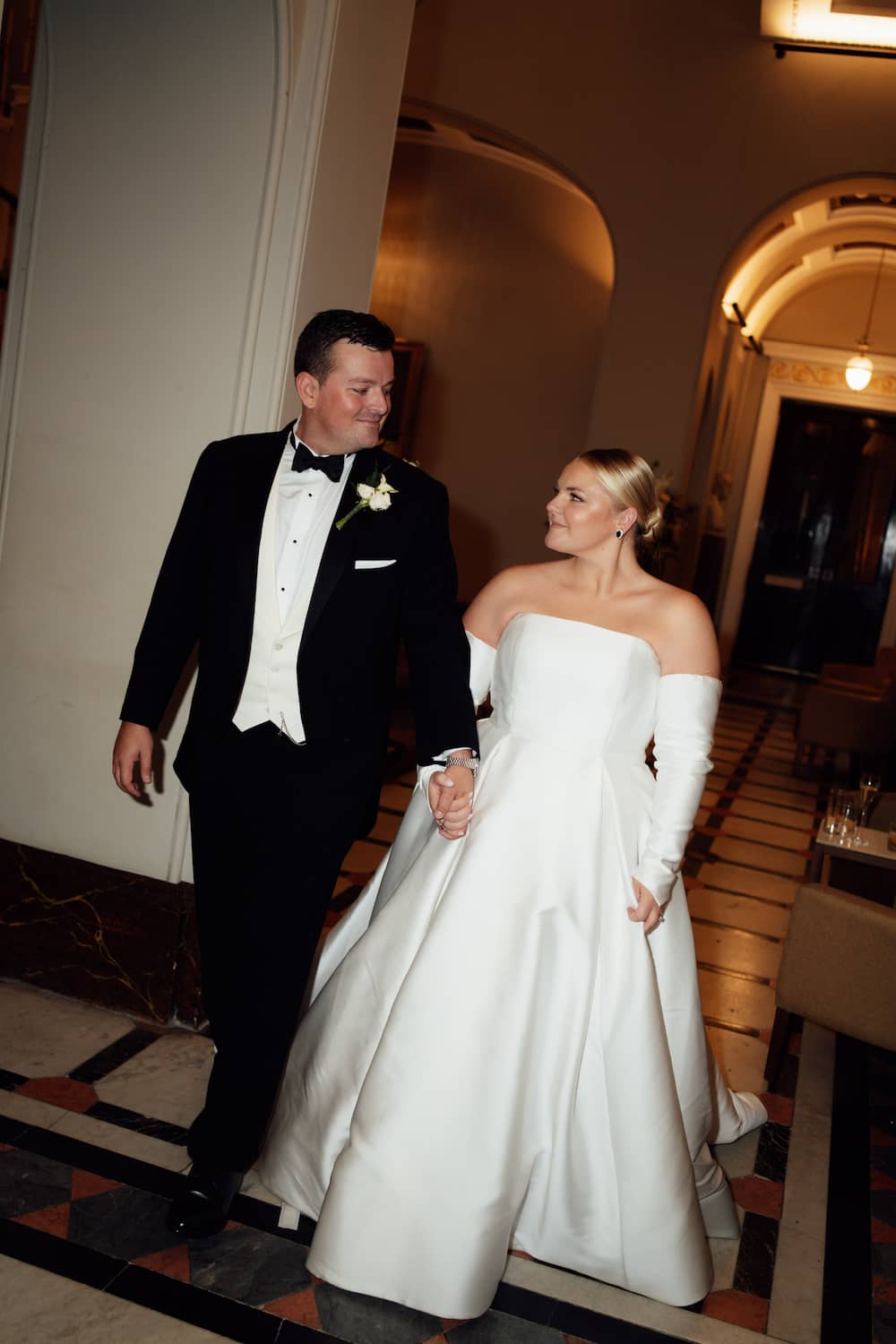 A bride in a strapless white satin wedding gown with long gloves walks hand-in-hand with a groom in a black tuxedo with a white vest and bow tie inside a warmly lit hotel hallway. Unedited.