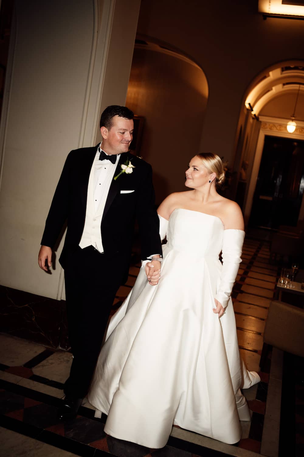 A bride in a strapless white wedding gown with long sleeves walks hand in hand with a groom in a black tuxedo with a white vest and bow tie down an indoor hallway, edited with Union of Two 2.0.