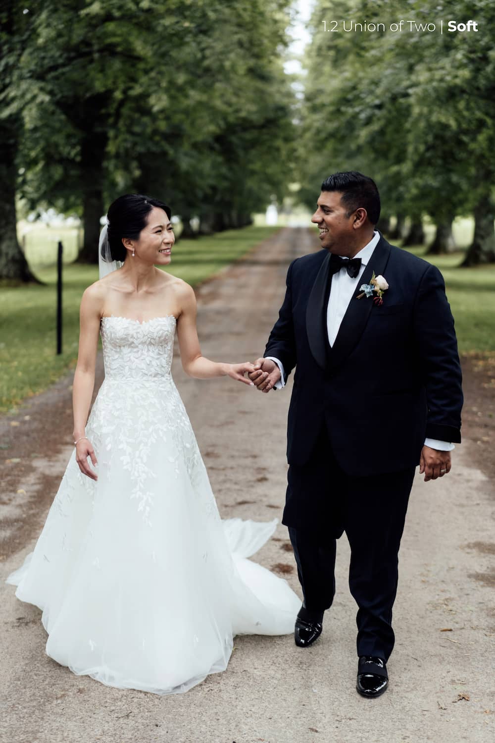 A bride in a white wedding dress and veil looks at her groom in a navy blue tuxedo as they walk down a tree lined driveway, edited with the Union of Two 2.0 Preset Pack.
