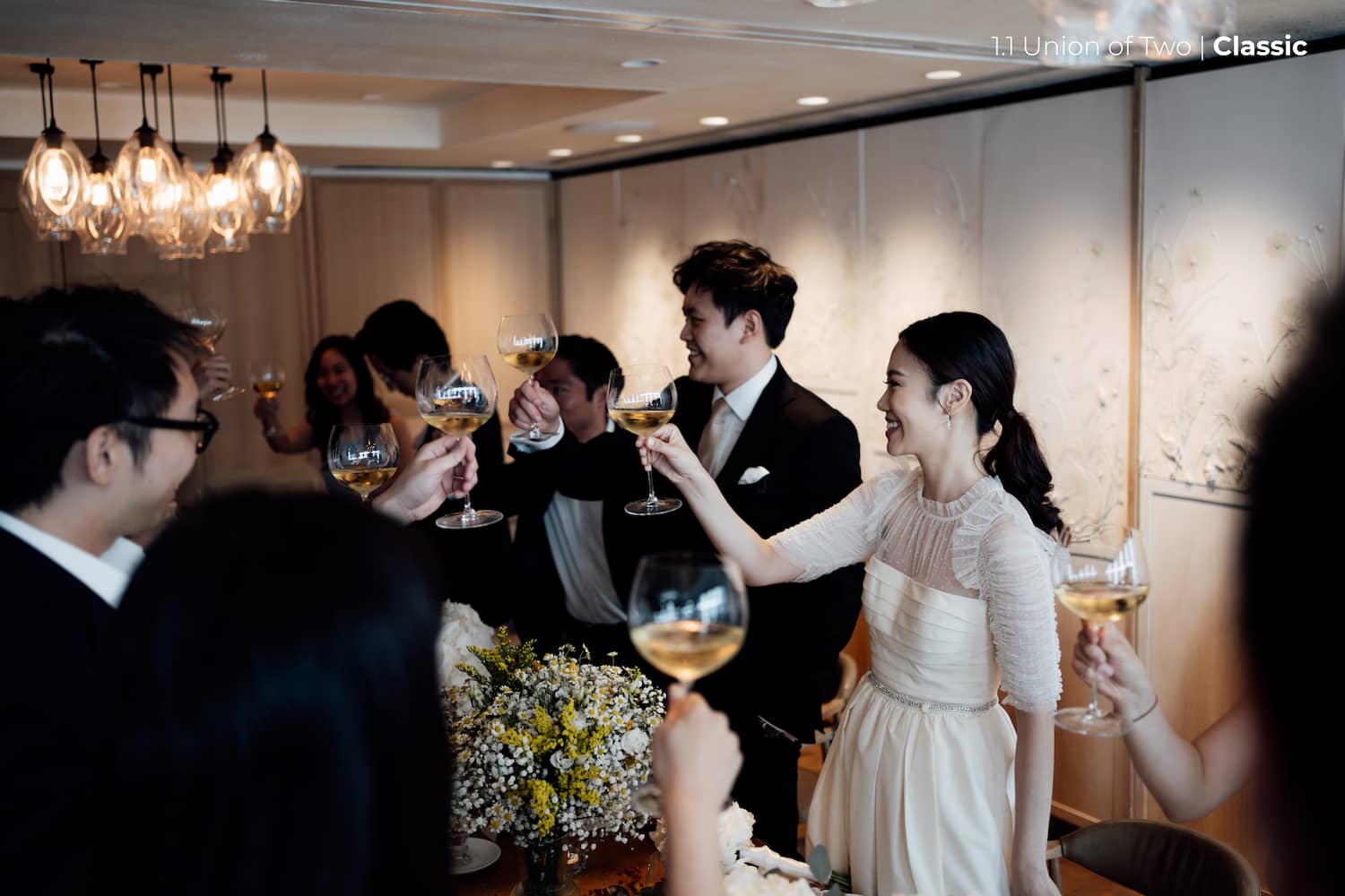A bride and a groom are performing a toast with their wedding party in a dining space with floral details, edited with the Union of Two 2.0 Preset Pack.