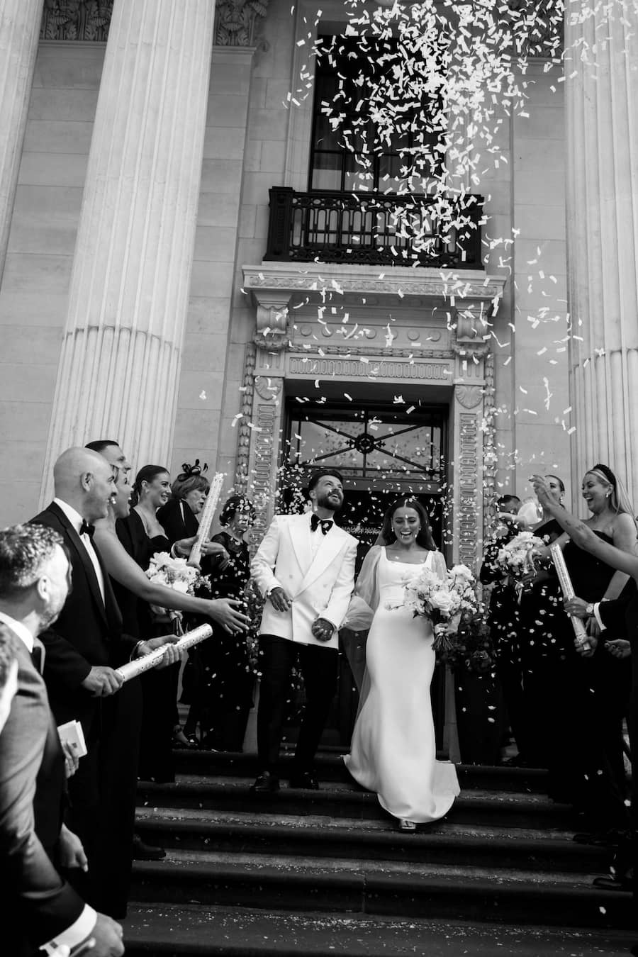 A wedding couple walking down stairs while guests through confetti over them, image is edited in black and white.