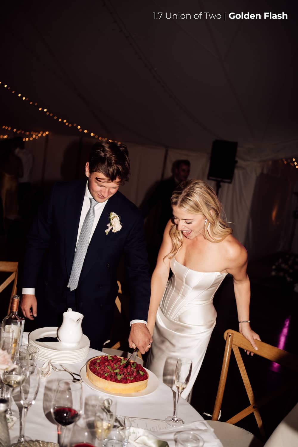 A bride and groom in wedding attire cut their wedding cake at a dining table, inside a marquee with lighting details, edited with the Union of Two 2.0 Preset Pack.