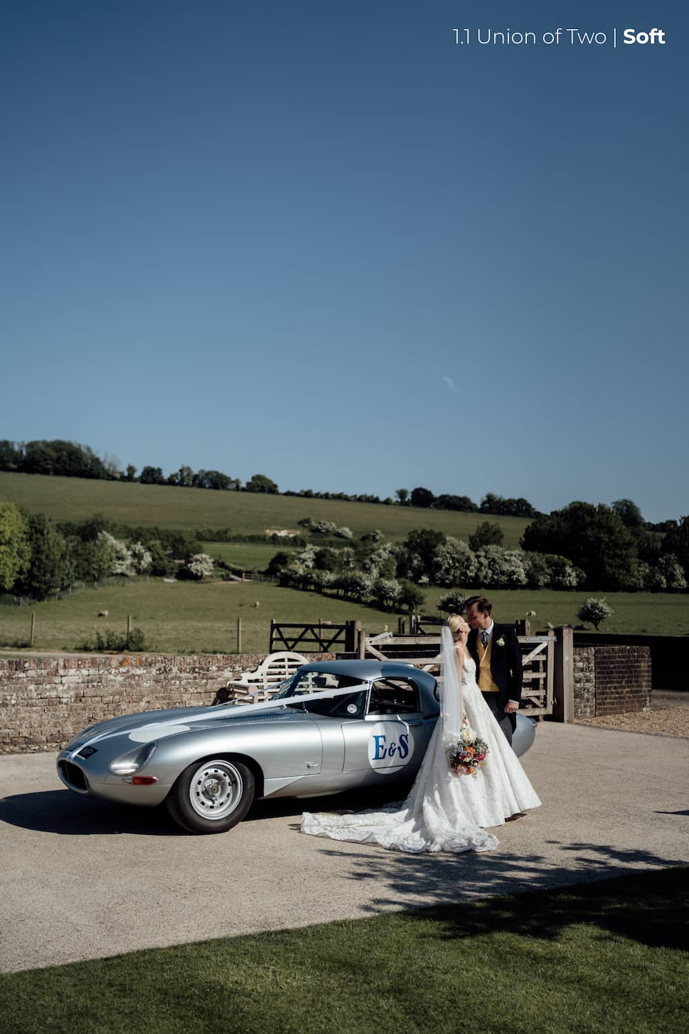 A bride in a long white wedding dress with a veil and holding a colourful bouquet of flowers faces her groom wearing traditional morning dress with a yellow waist coat. Behind them is a vintage jaguar e type sports car, in a countryside scene with rolling hills,  edited with the Union of Two 2.0 Preset Pack.