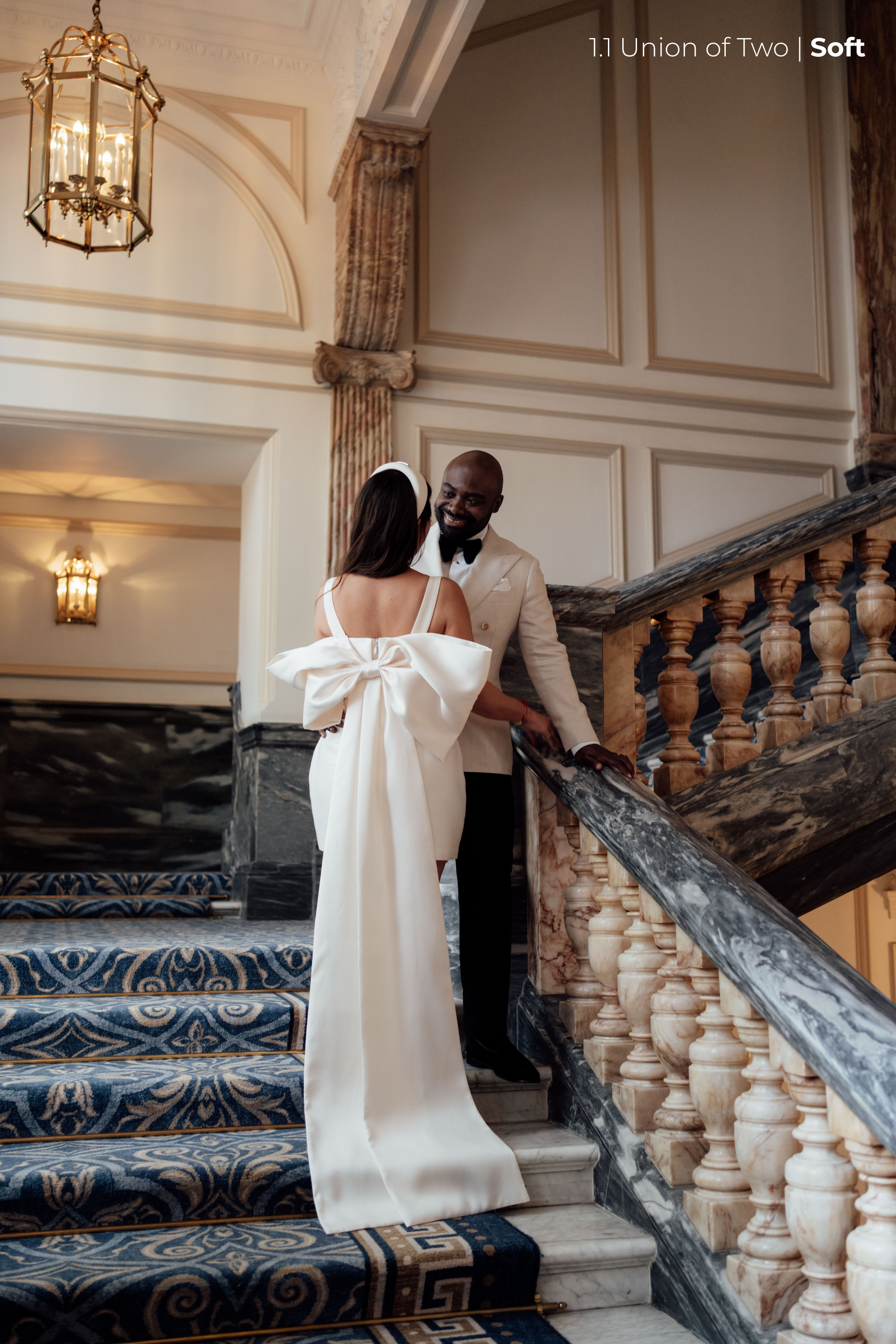 A bride in a white dress with a long bow detail and a white headband, with a groom in a white tuxedo are standing on a staircase,  edited with Union of Two 2.0 Preset Pack.