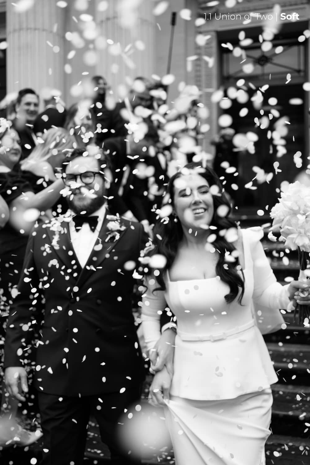 A bride in a short white wedding dress and a groom in glasses and a black tuxedo are having confetti thrown over them, edited with Union of Two 2.0 Preset Pack.