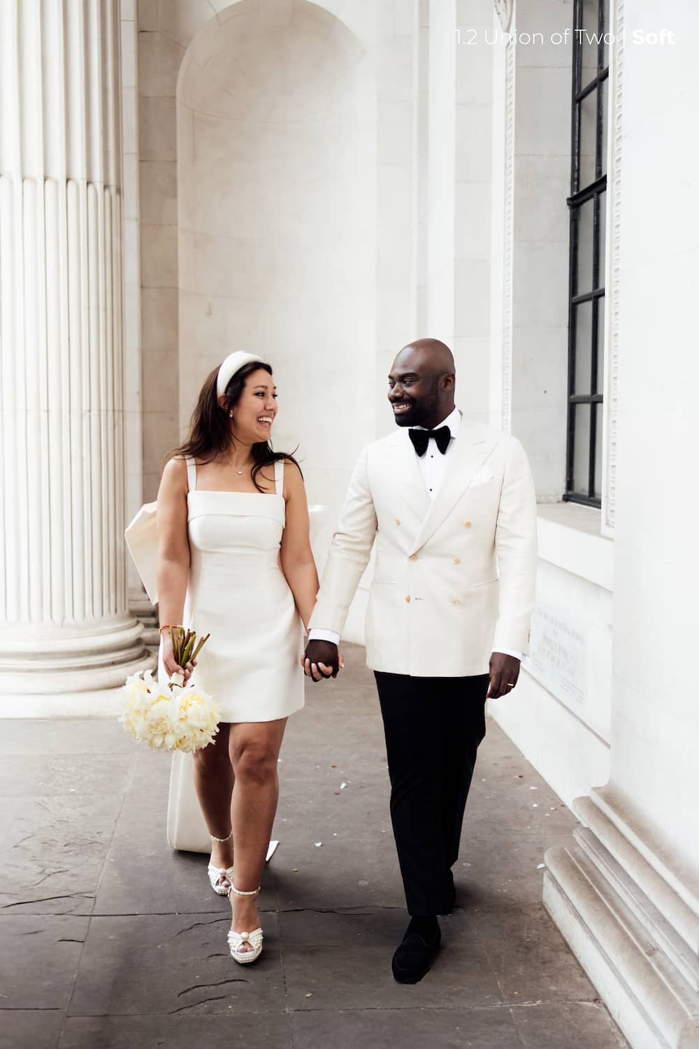 A bride in a short white wedding dress holding a white bouquet of flowers and a groom in a white tuxedo walking towards the camera, edited with Union of Two 2.0 Preset Pack.