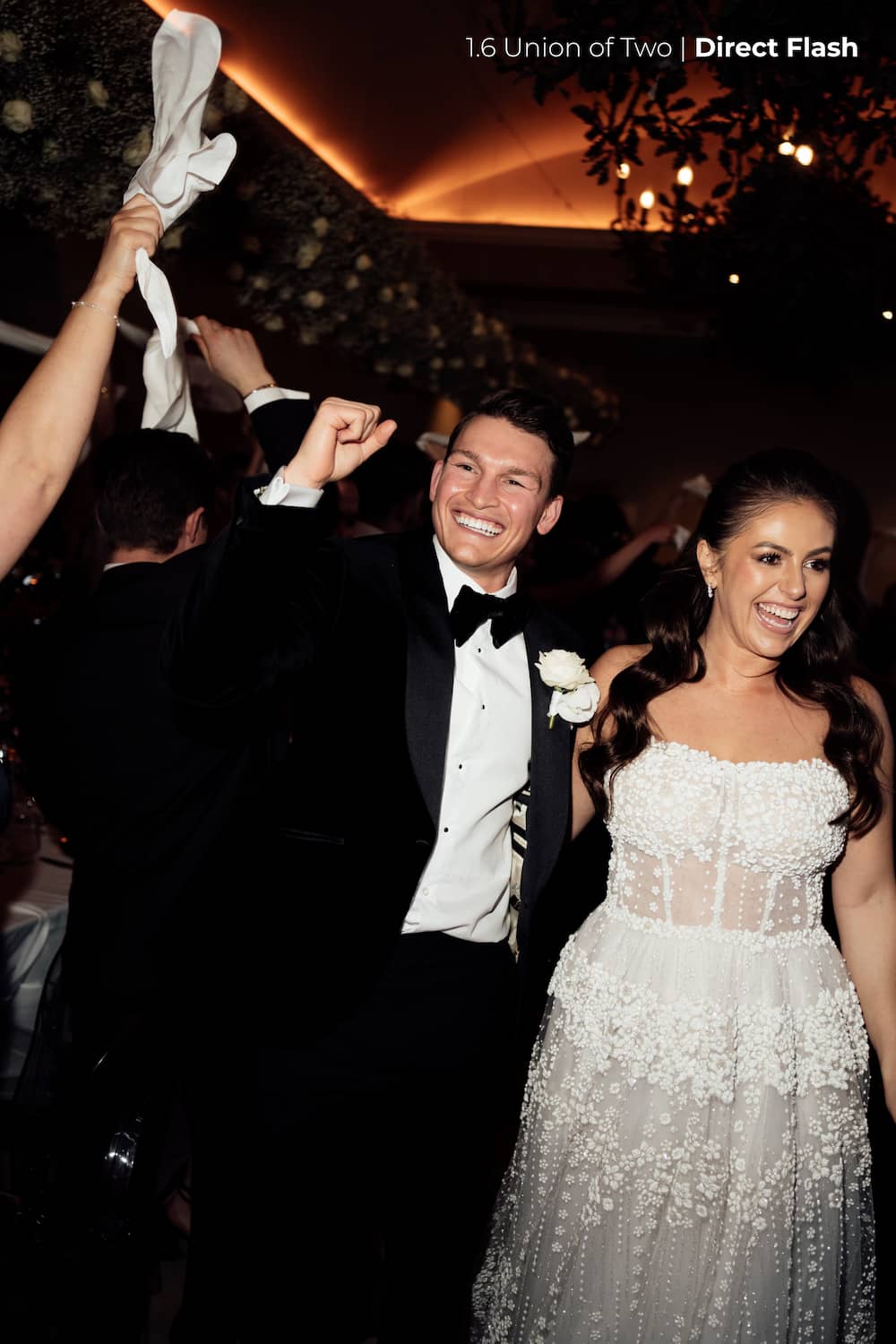 A bride in a white wedding dress and groom in a black tuxedo celebrating as they enter a room during their wedding edited with Union of Two 2.0 Preset Pack.