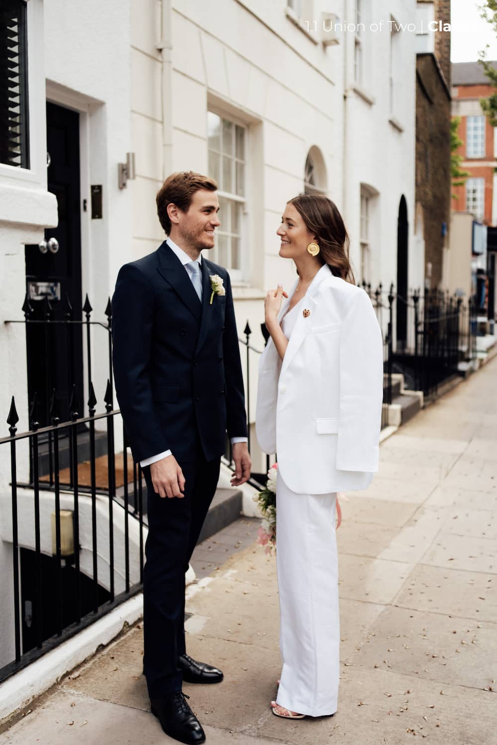 A bride and groom standing on a London street. The groom is wearing a dark navy suit with a white shirt and tie, and the bride is wearing a matching white pants suit with a white blazer, edited with the Union of Two 2.0 Preset Pack.