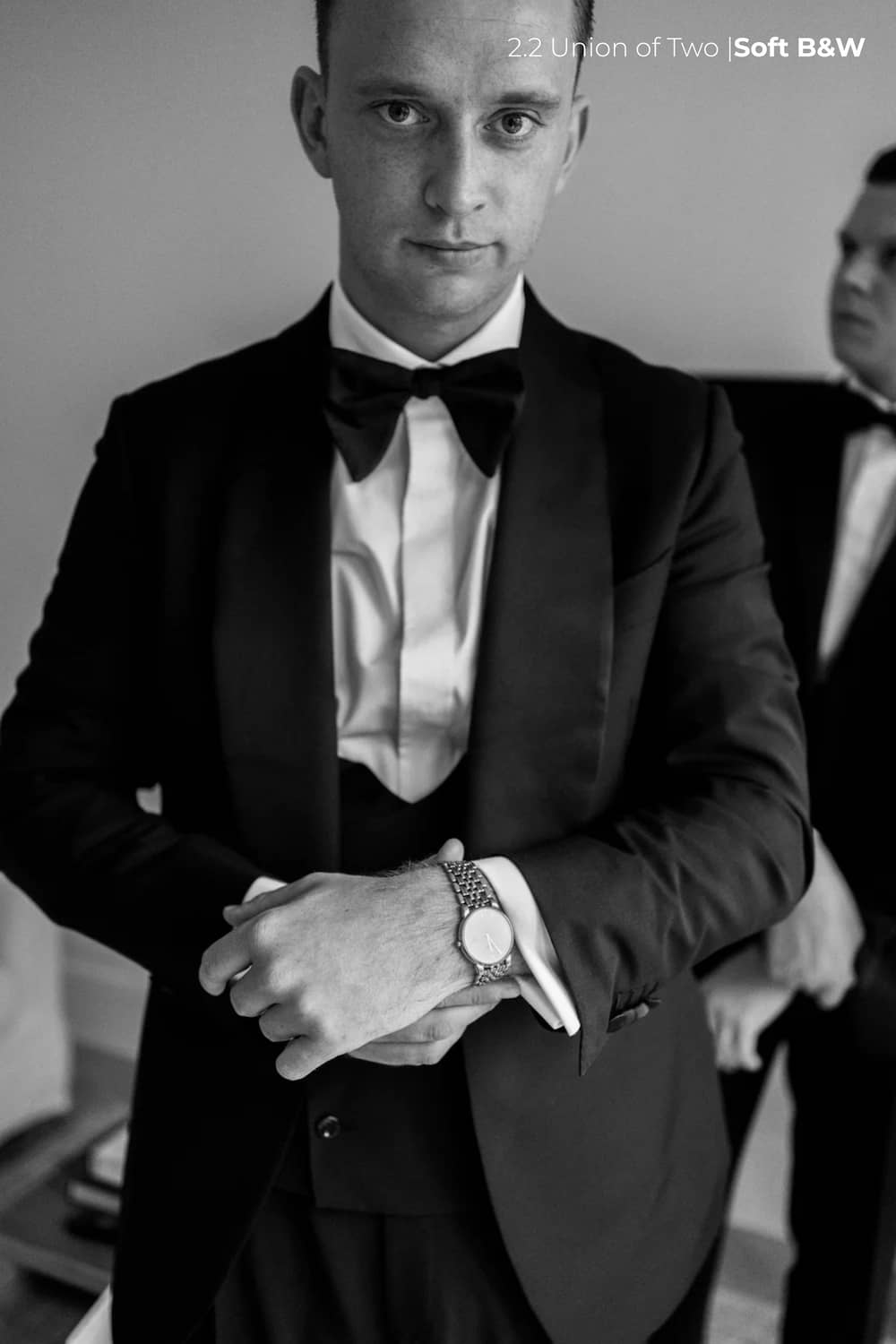 A groom in a black tuxedo adjusts his watch, with a groomsman visible in the background, edited in black and white with the Union of Two 2.0 Preset Pack.