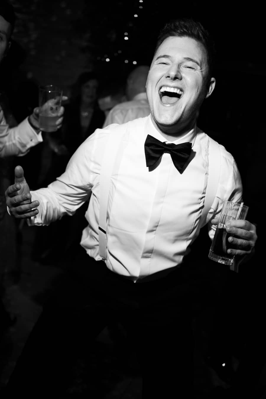 A man in a white dress shirt, black bow tie, and suspenders dancing and laughing at a night event, holding a drink.
