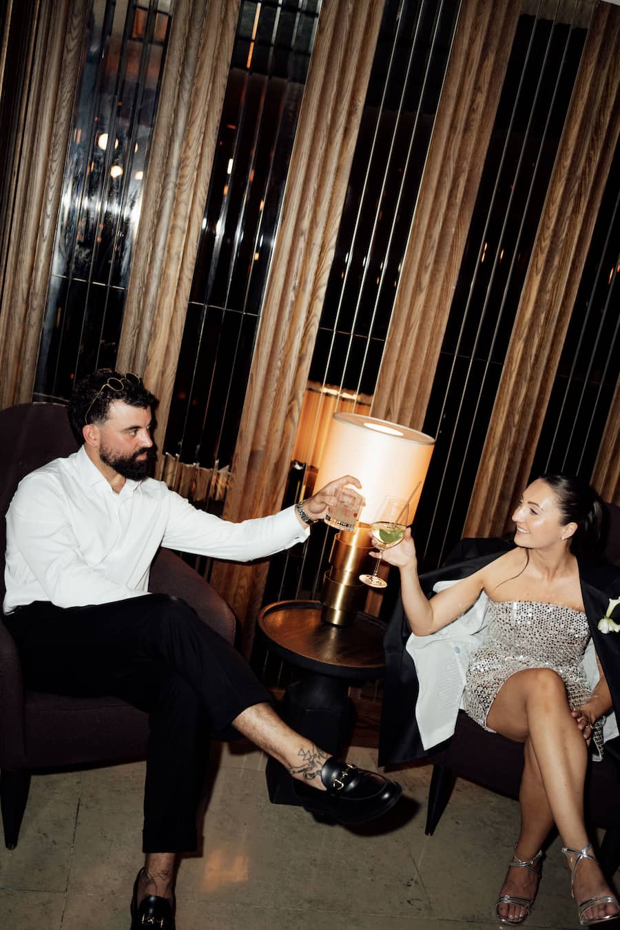 A man in a white shirt and black trousers and a woman in a silver strapless dress clinking glasses in a dimly lit lounge setting with a lamp between them.