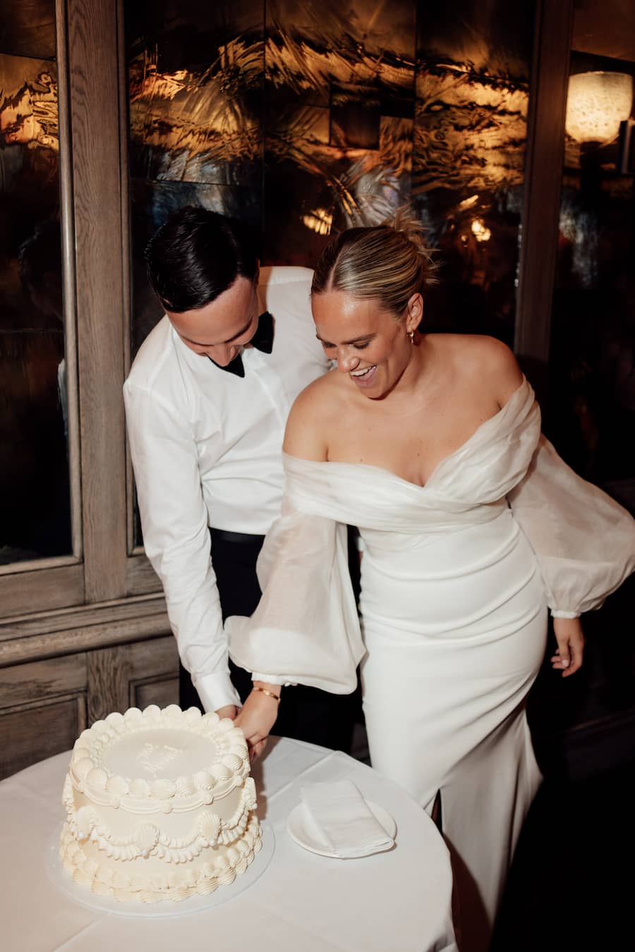 A bride in an off-the-shoulder white gown and a groom in a white shirt and black bow tie cut a white frosted wedding cake together in a warmly lit indoor setting.