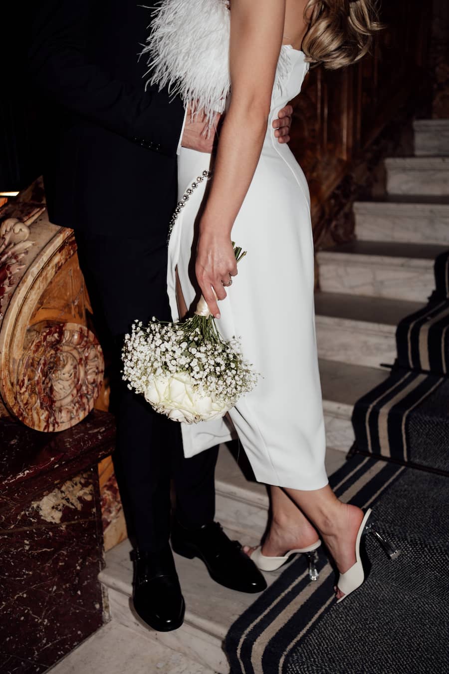 A bride in a strapless white wedding dress with a white feather boa and holding a bouquet of baby's breath, walking down a staircase next to a groom in a black suit.