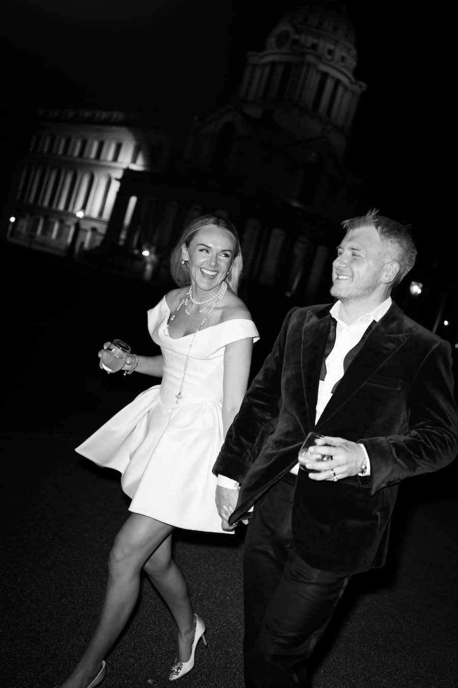 A black and white photograph of a smiling couple walking hand-in-hand at night in front of a grand illuminated building. The woman wears an off-the-shoulder white mini dress, and the man wears a dark velvet jacket and white shirt.