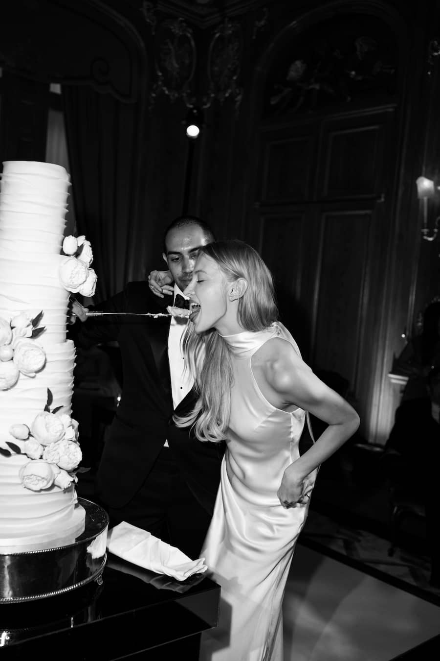 Black and white photograph of a couple sharing a large tiered wedding cake with a knife, with the woman in a satin sleeveless gown leaning in to take a bite.