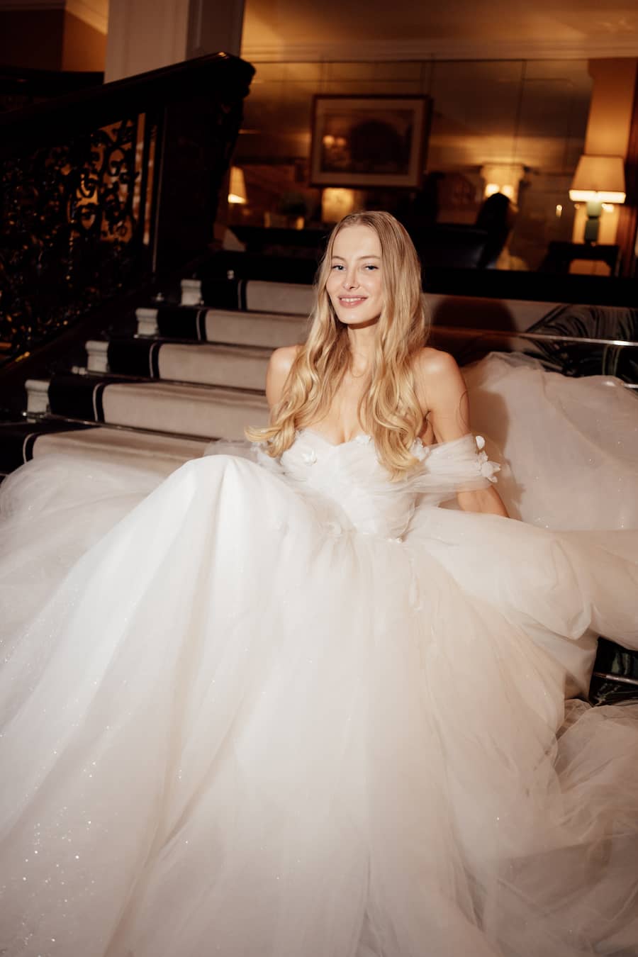 A woman in a voluminous white off-the-shoulder wedding gown sits on a grand staircase in a warmly lit interior.
