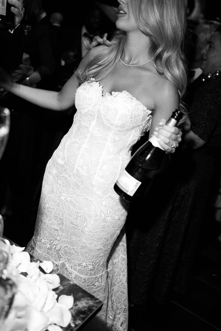 A woman in a strapless lace wedding gown holding a champagne bottle in a dark, crowded indoor setting. The photo is in black and white.