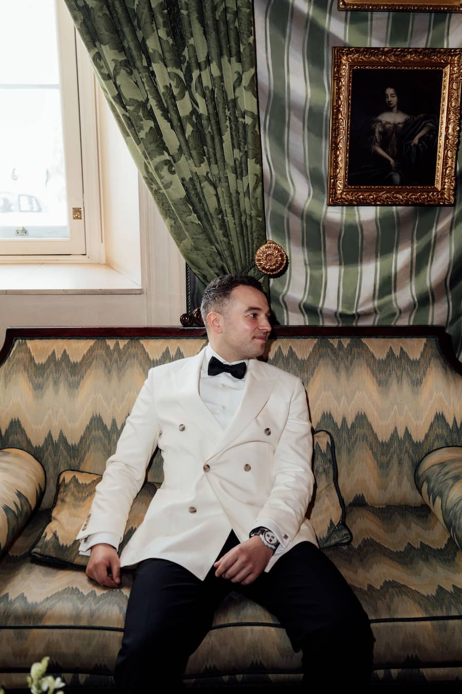 A man wearing a white double-breasted tuxedo jacket with gold buttons, a white shirt, and a black bow tie sits on a patterned sofa in an elegant room with green striped wallpaper and a window.