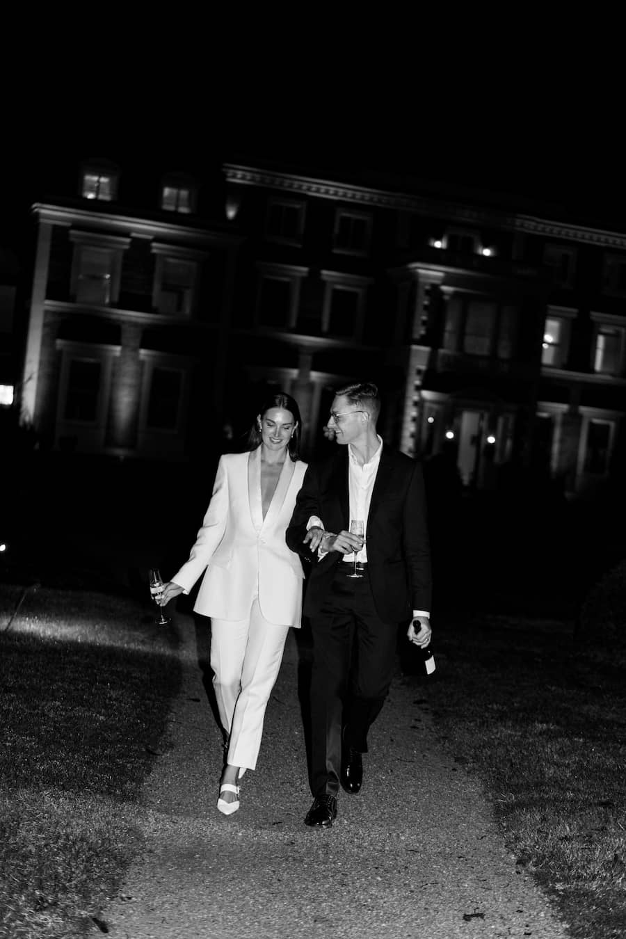 A black and white photograph of a couple walking arm-in-arm at night in front of a grand building. The woman is wearing a light-colored, tailored suit, and the man is in a dark suit.