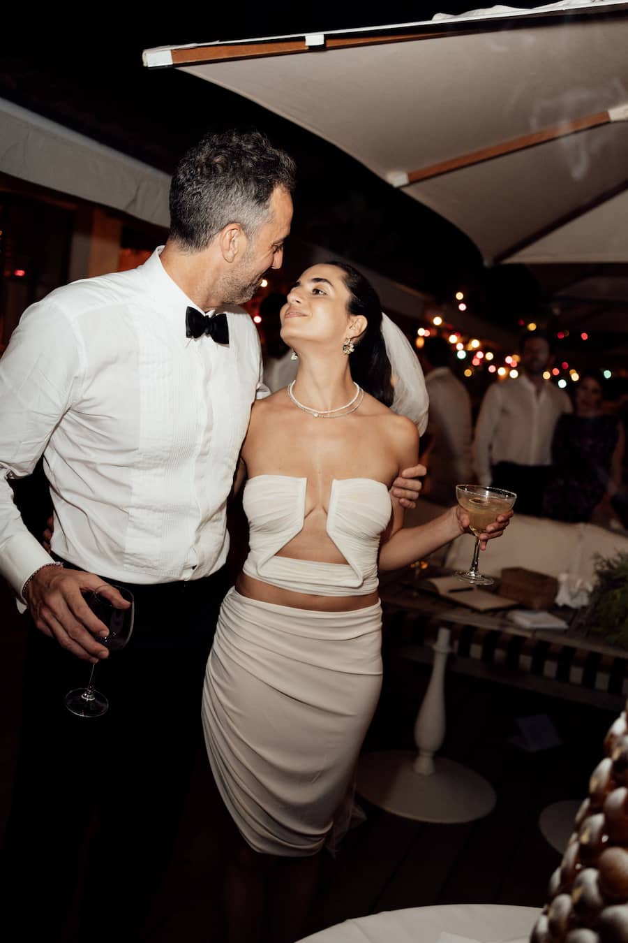 A couple embracing at a nighttime event. The woman wears a strapless cream-colored dress with a cutout bodice and a ruched skirt, while the man wears a white dress shirt and black bow tie.